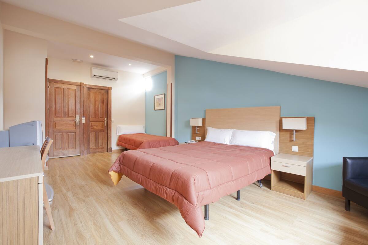 A hotel room with two beds, a desk, and a chair. There is a wooden wardrobe and a nightstand with a lamp.
