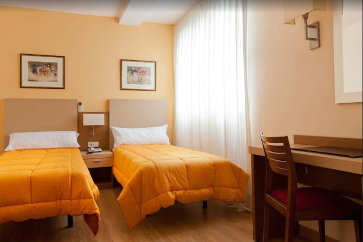 A hotel room with two beds, a desk, and a chair. There are two framed pictures on the wall.