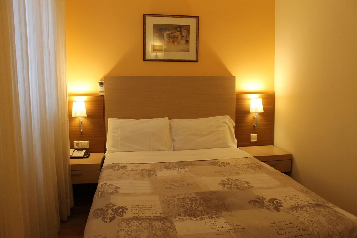 A hotel room with a bed, two bedside lamps, a nightstand with a phone, and a framed picture on the wall.