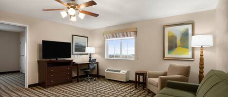 A hotel room with a ceiling fan, a flat-screen TV, a desk with a chair, a sofa, a window with a view, and a painting on the wall.