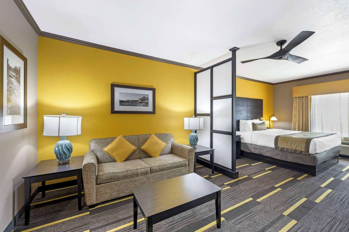 A hotel room with a yellow accent wall, a sofa, a bed, a ceiling fan, and a large window with curtains.