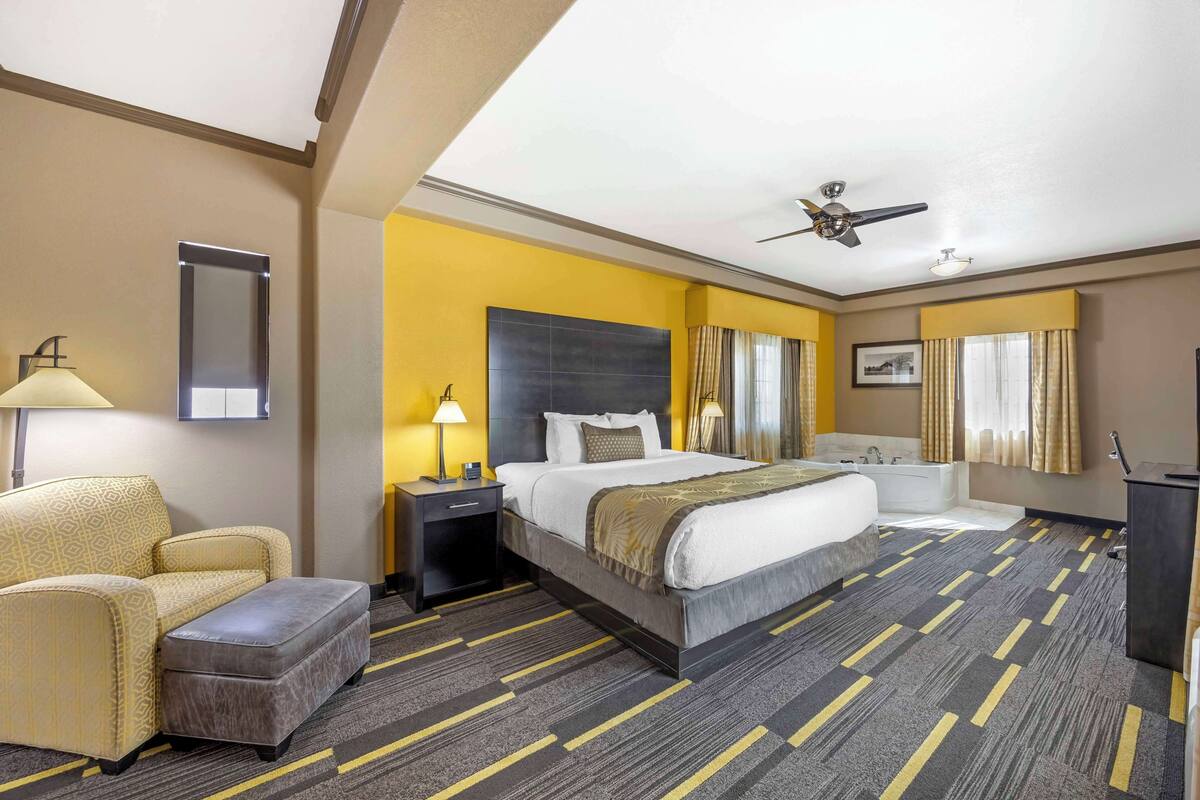 A hotel room with a bed, a sofa, a desk, a lamp, a mirror, a ceiling fan, and a window with curtains.