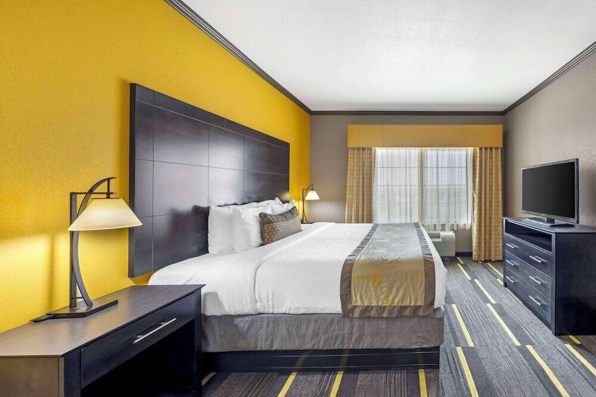 A hotel room with a large bed, a flat-screen TV, a desk with a lamp, and a window with curtains.