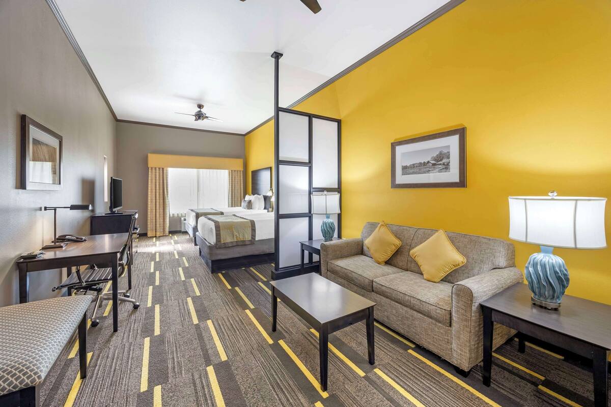 A hotel room with a sofa, a coffee table, a desk, a chair, a TV, a bed, and a window with curtains.