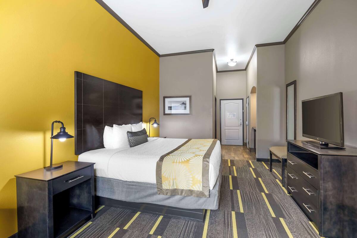 A hotel room with a bed, bedside table, lamp, TV, and a door leading to a hallway.