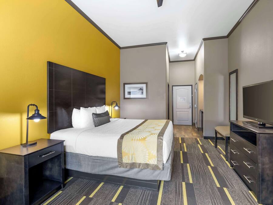 A hotel room with a bed, bedside table, lamp, TV, and a door leading to a hallway.