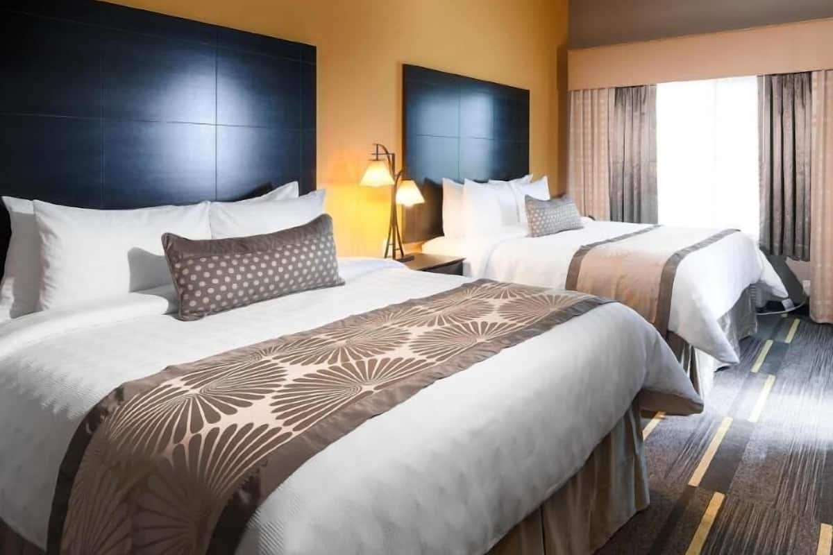 A hotel room with two beds, a headboard, a nightstand, a lamp, and a window with curtains.