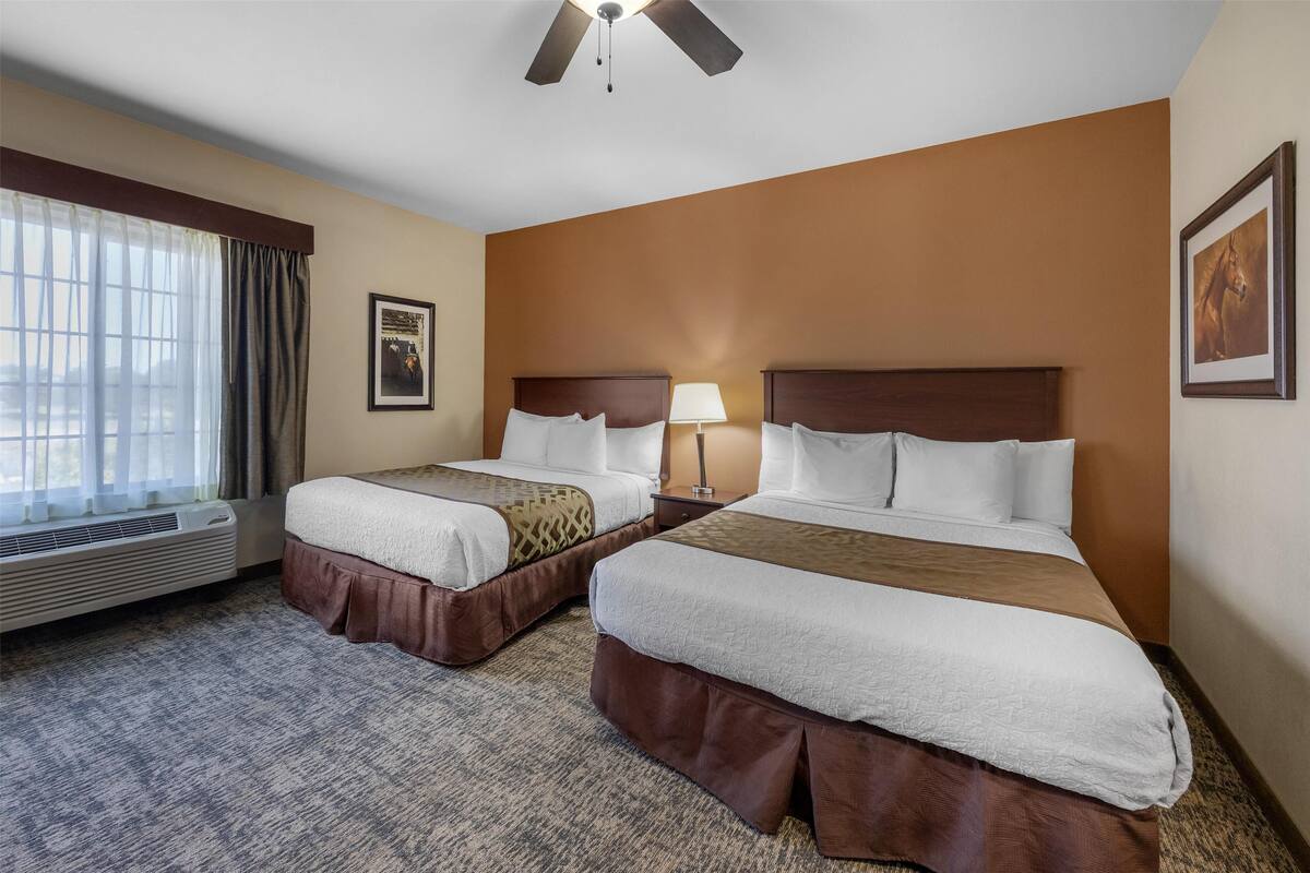 A hotel room with two beds, a ceiling fan, a window with curtains, and a framed picture on the wall.