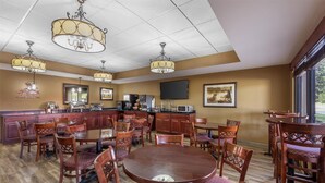 A spacious dining area with wooden tables and chairs, a kitchen in the background, and large windows. - Best Western North Edge Inn (Dodge City)