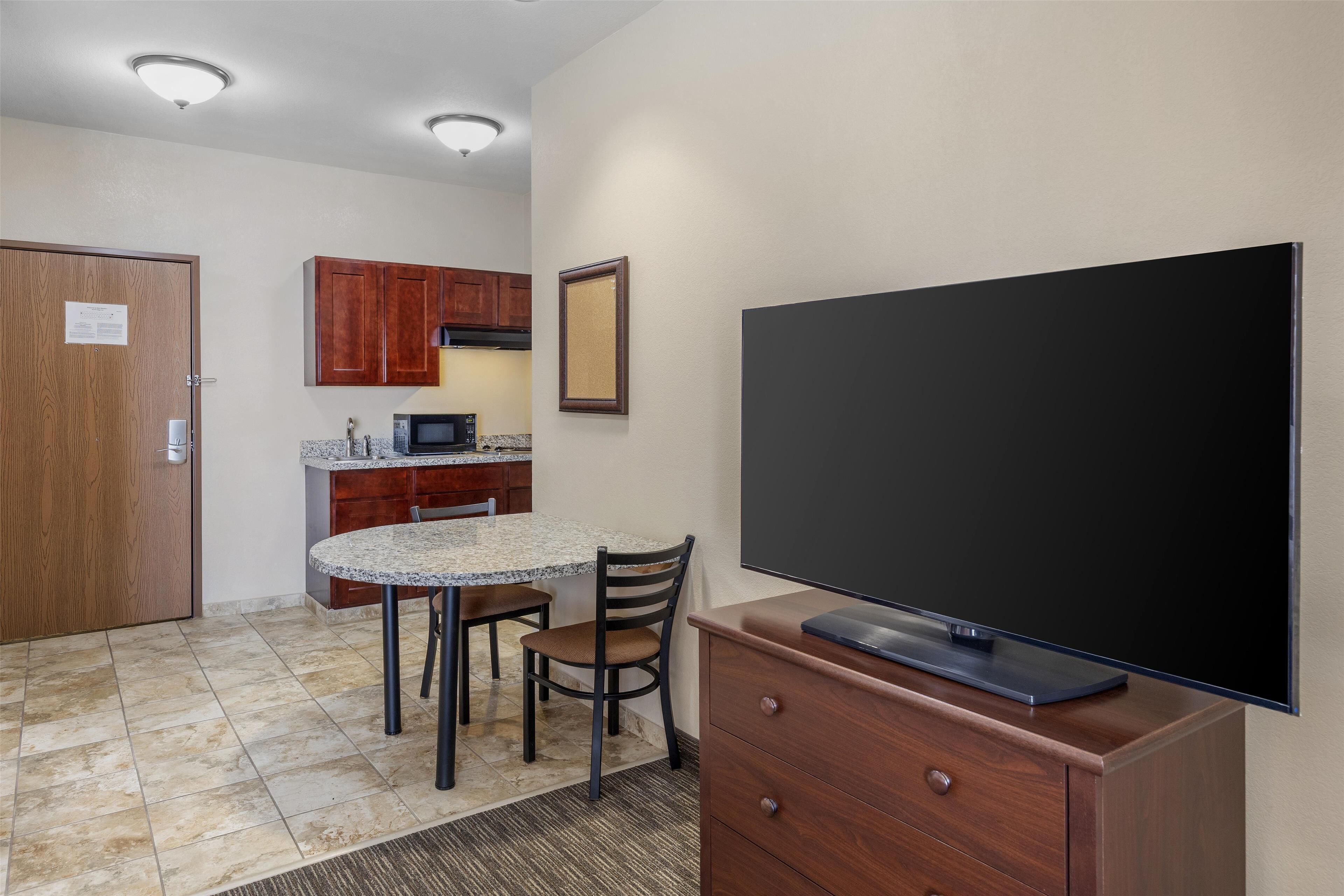A compact kitchen with a granite countertop, wooden cabinets, a microwave, and a flat-screen TV on a wooden stand.