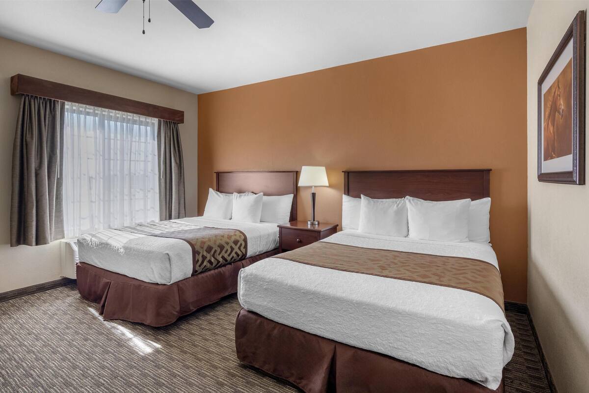 A hotel room with two beds, a nightstand with a lamp, a large window with curtains, and a ceiling fan.