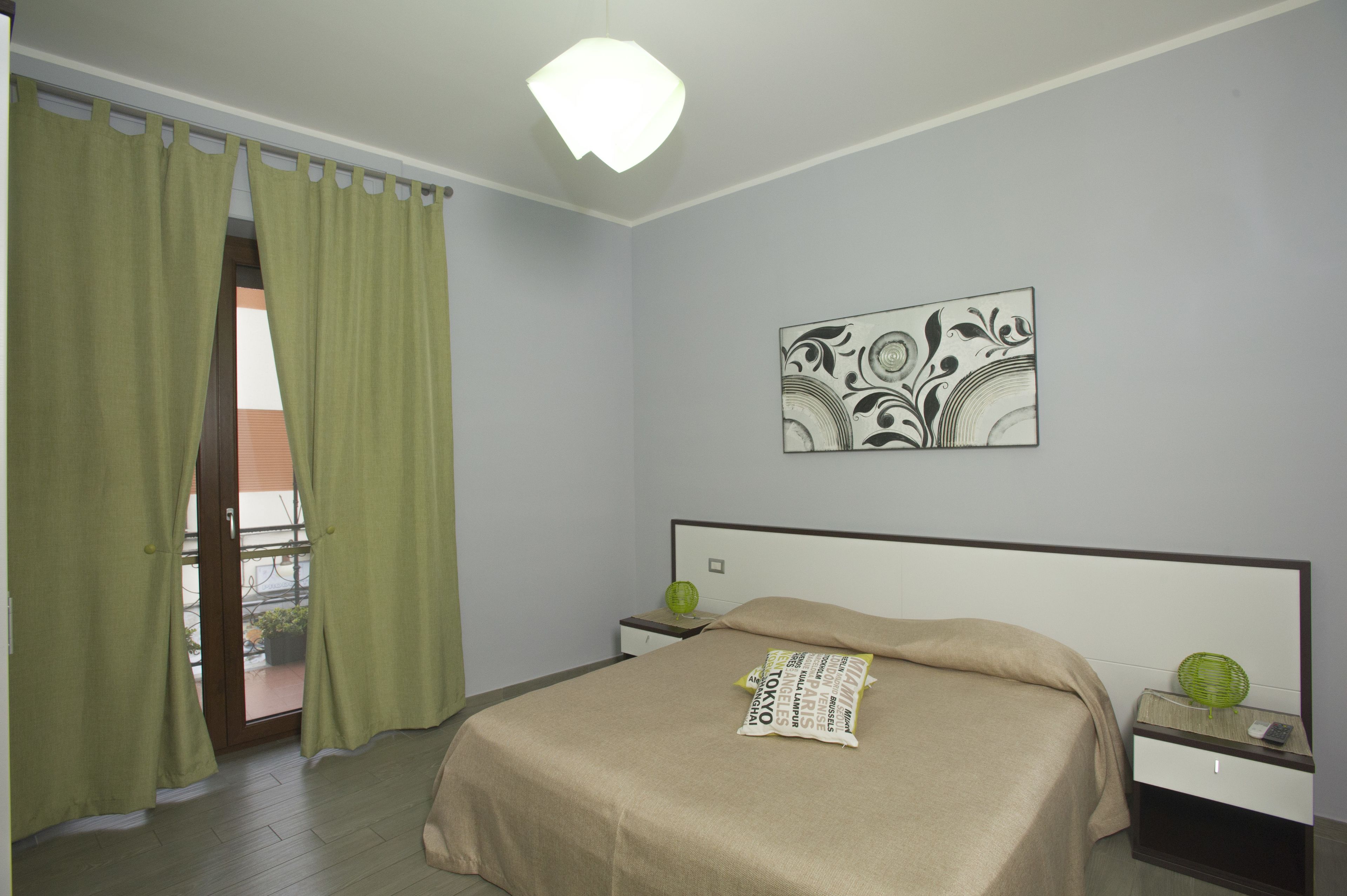 A hotel room with a bed, a nightstand, a wall-mounted artwork, and a view through a window with curtains.