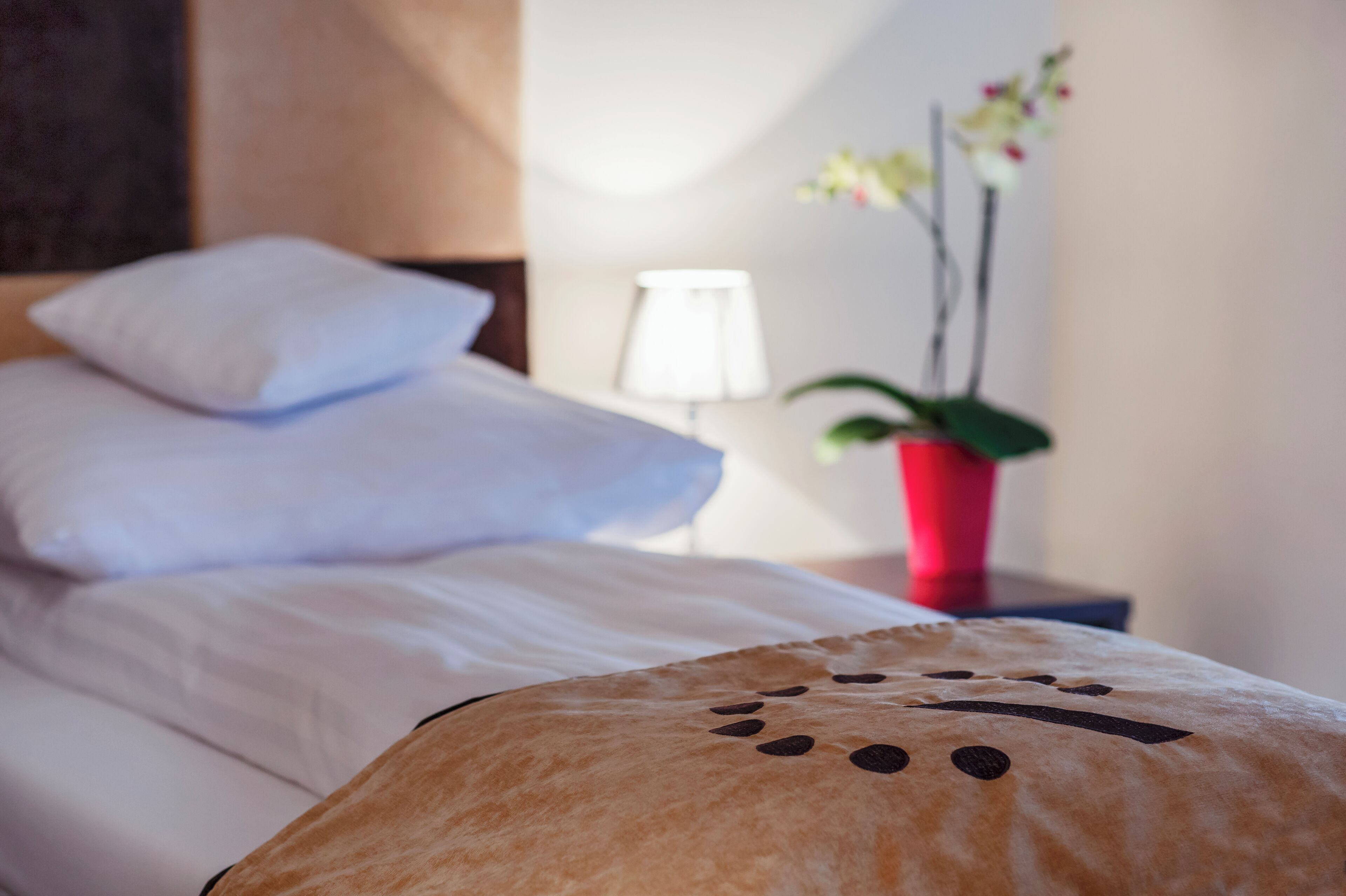 Una cama bien hecha con sábanas blancas, una lámpara de noche y un jarrón rojo con flores.