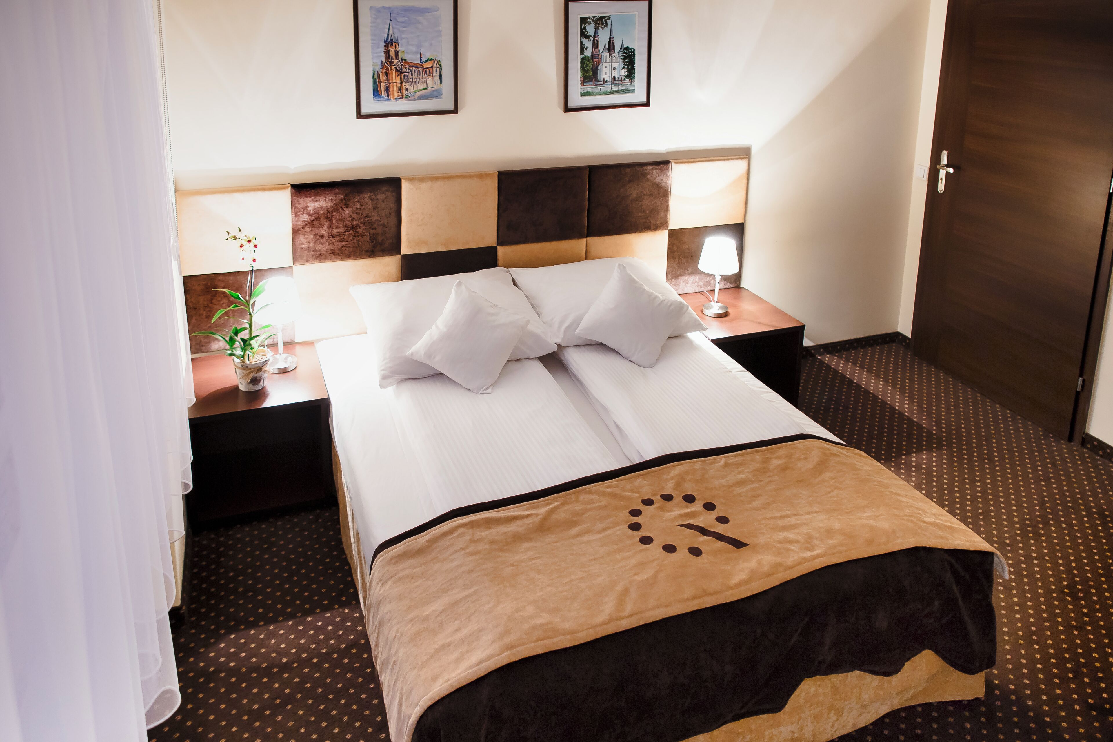 A hotel room with a neatly made bed, a nightstand with a lamp, and two framed pictures on the wall.