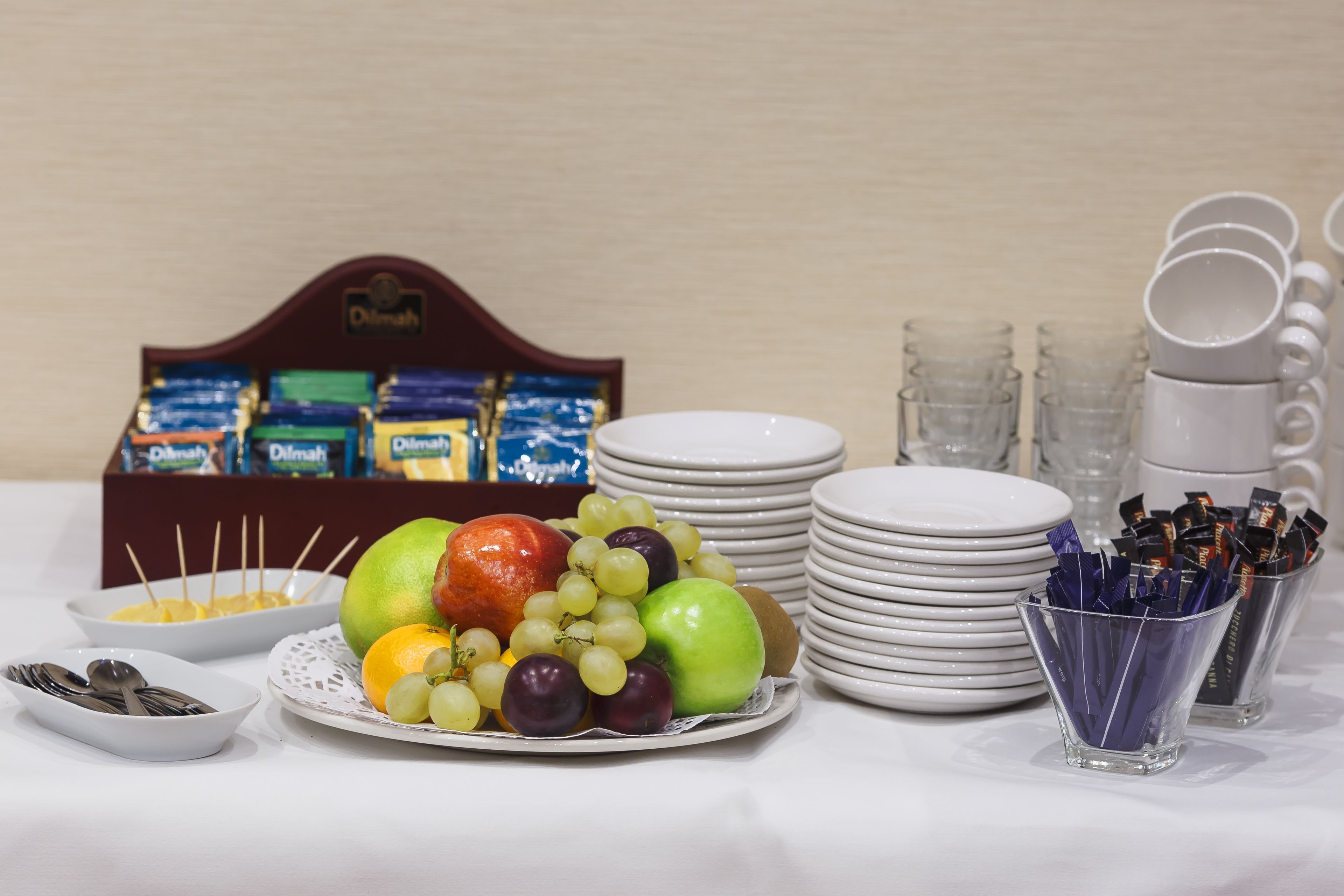 Una mesa con una bandeja de frutas, platos, vasos y una caja de bombones.
