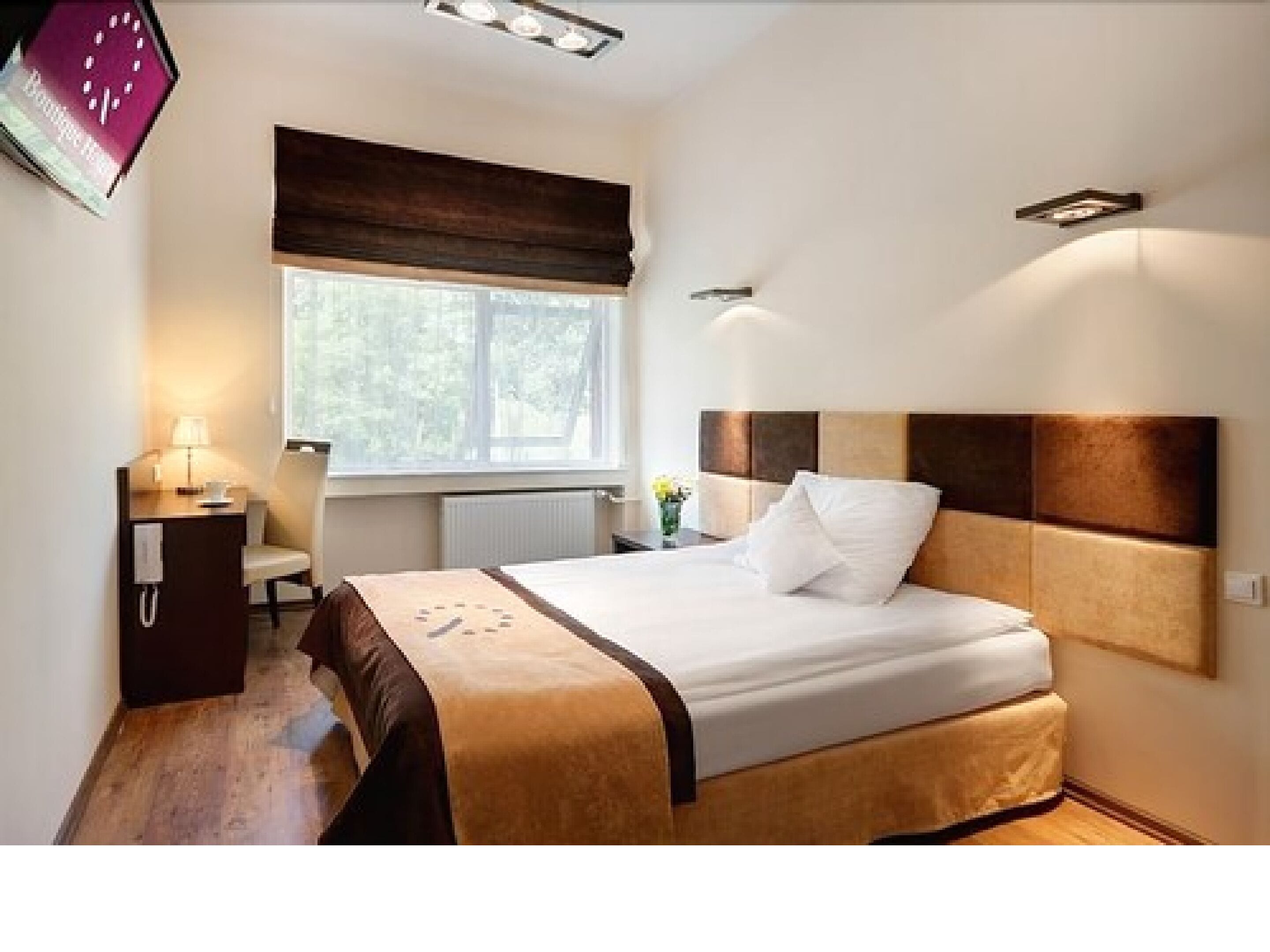 Una habitación de hotel con cama, mesita de noche, lámpara y ventana con vistas a árboles.