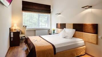 Una habitación de hotel con cama, mesita de noche, lámpara y ventana con vistas a árboles.