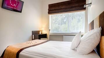 A hotel room with a bed, a bedside lamp, a window with a view of trees, and a wall-mounted TV displaying the hotel name.