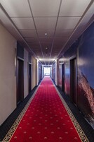 Um corredor comprido e estreito com carpete vermelho, portas em ambos os lados e teto com iluminação embutida.