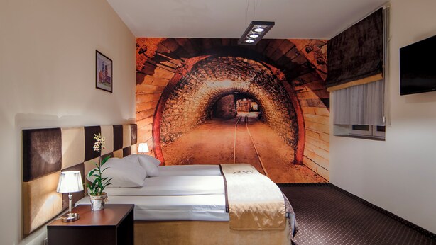 Habitació d'hotel amb un llit, una tauleta de nit amb una làmpada i una planta, un mural a la paret que representa un túnel i una finestra amb cortines.