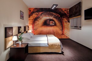 Quarto de hotel com cama, mesa de cabeceira com abajur e planta, painel na parede representando um túnel e janela com cortinas.