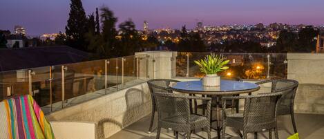 Una terraza en la azotea con una mesa redonda y sillas, una planta en maceta y vistas a un paisaje urbano al atardecer.