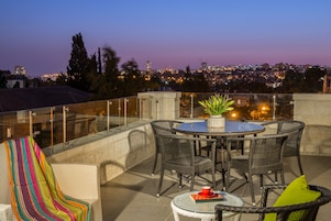 A rooftop terrace with a round table and chairs, a potted plant, and a view of a cityscape at dusk.