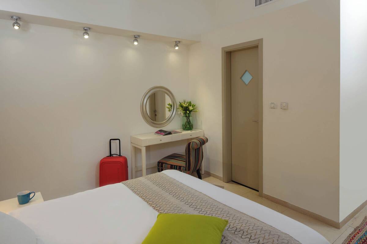 Een hotelkamer met een bed, een bureau met spiegel, een rode koffer en een deur.