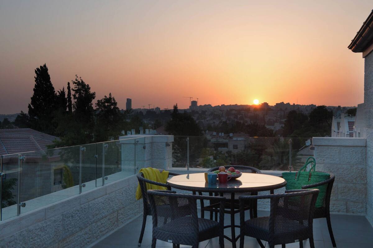 Een ronde tafel met stoelen op een balkon met uitzicht op een stadsbeeld bij zonsondergang.