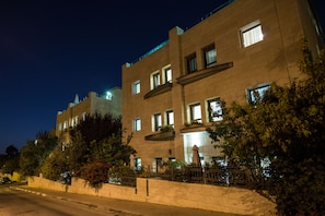 A multi-story building with illuminated windows and balconies, surrounded by greenery and a paved sidewalk.