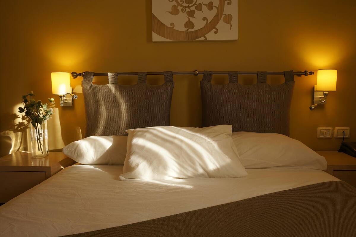Een net opgemaakt bed met witte lakens en twee kussens, een nachtkastje met een bloevas, en twee wandlampen.