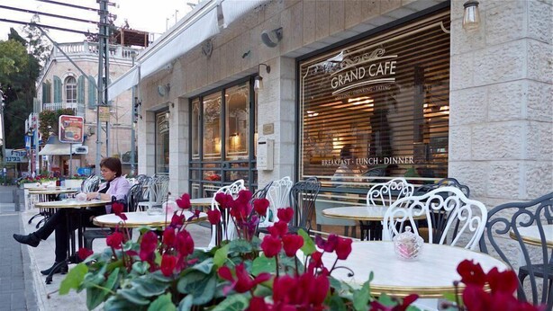 Un café en plein air avec des tables et des chaises, une femme assise à une table et un bâtiment sur lequel on peut lire l’inscription « Grand Café ».