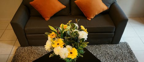 A living room with a dark grey sofa, two orange cushions, and a black coffee table with a floral arrangement.