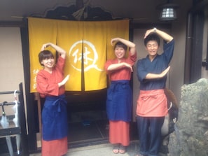 A group of people in traditional attire standing in front of a yellow curtain with a white symbol. - Gion Ryokan Q-beh - Hostel (Kyoto)