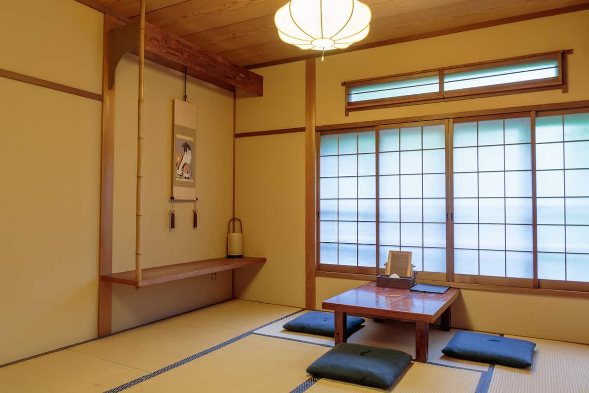 A traditional Japanese room with tatami flooring, a low wooden table, cushions, a hanging lantern, and a shoji screen window.