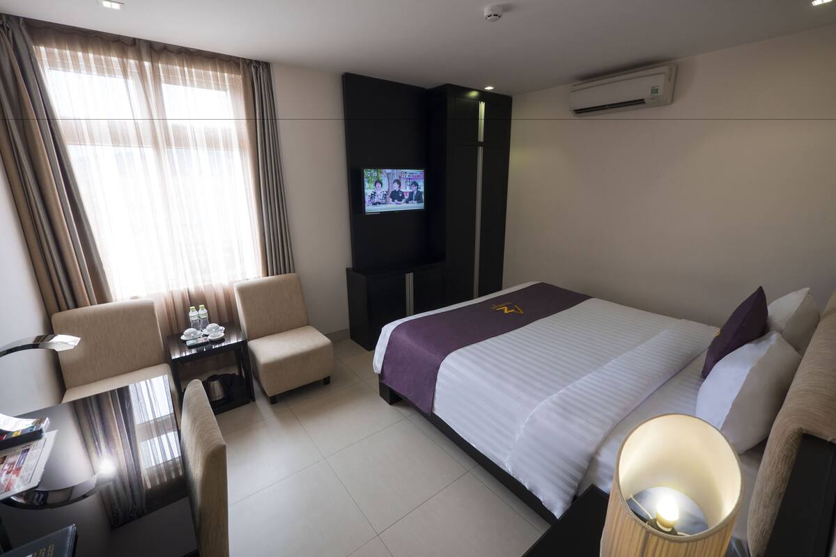 A hotel room with a bed, a desk, a chair, a TV, and an air conditioning unit.