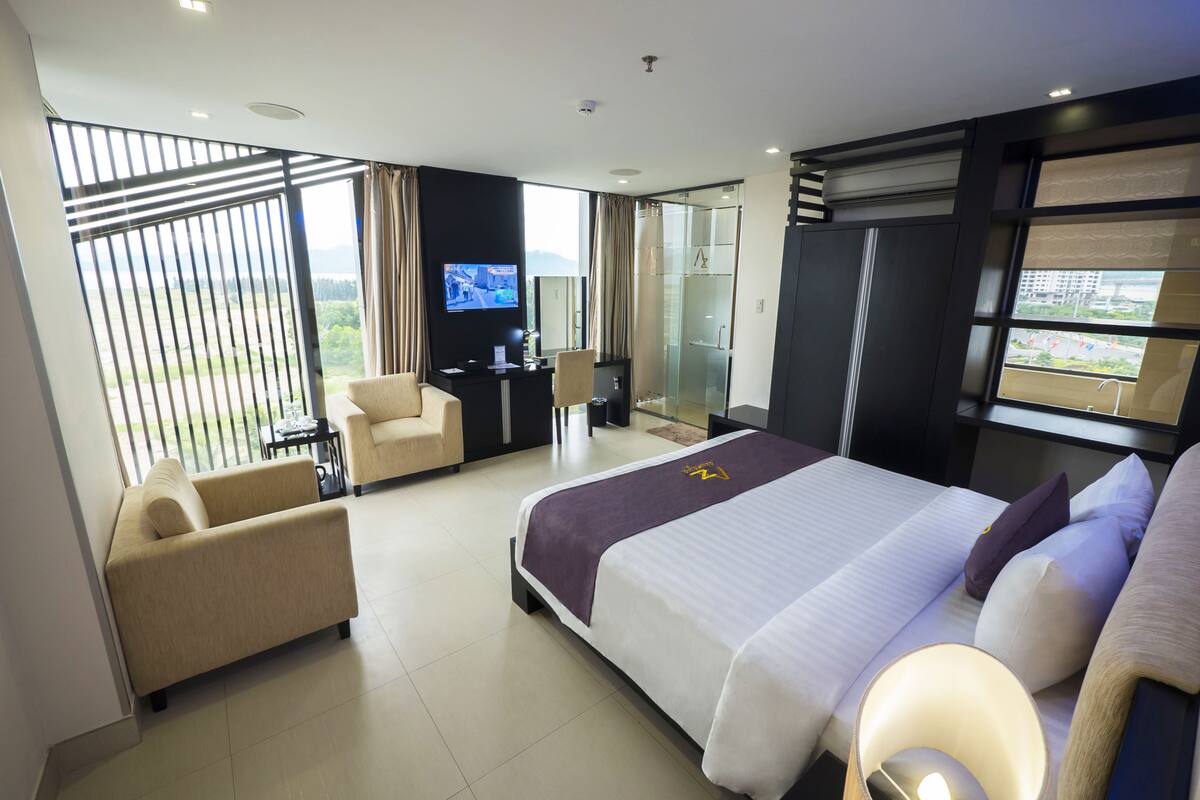 A modern hotel room with a large bed, a sofa, a desk with a TV, and a glass shower area.