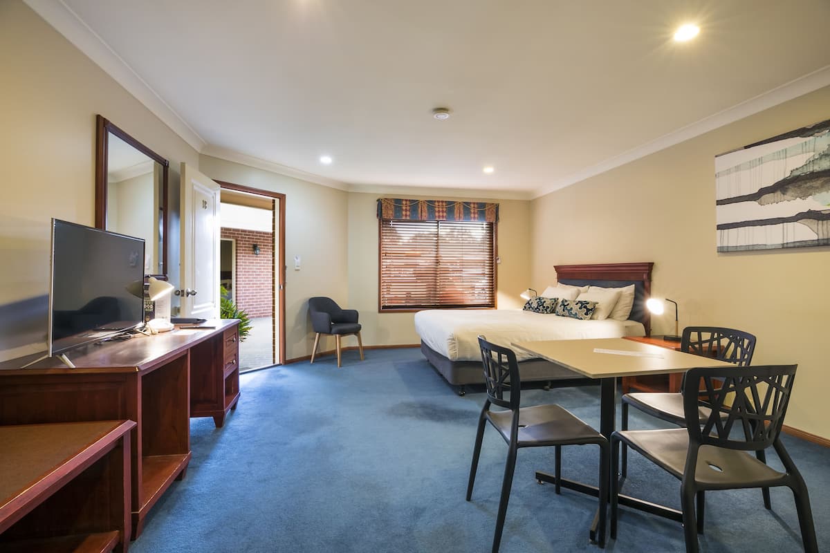 A hotel room with a bed, desk, chairs, a TV, and a window with blinds.