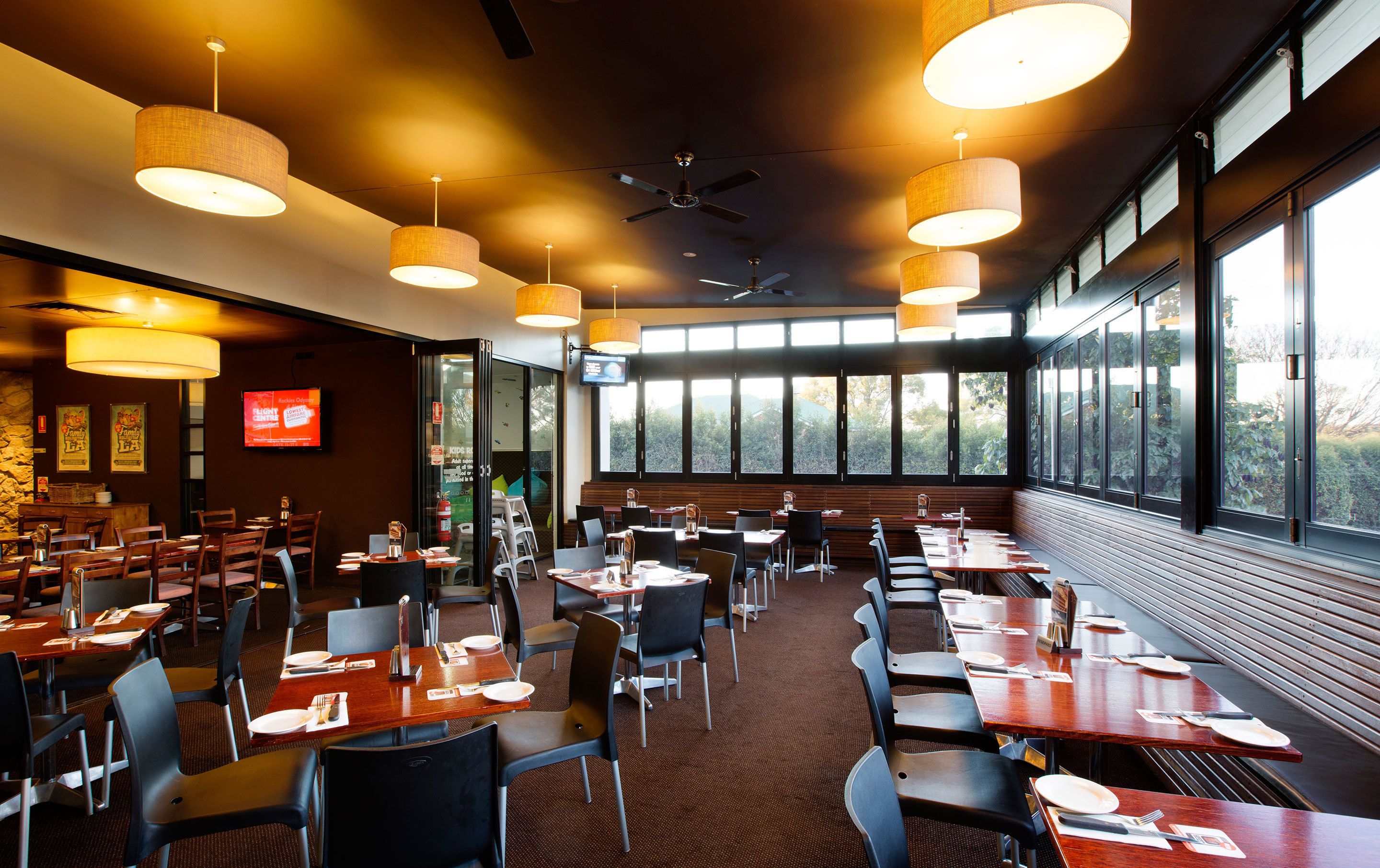 Un restaurante con varias mesas y sillas, amplias ventanas con vistas, ventiladores de techo y lámparas colgantes.