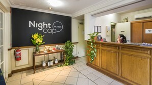 A reception area with a black wall displaying "Nightcap Hotels" logo, a wooden reception desk, a fire extinguisher, a scale, and a plant. - Nightcap at Federal Hotel Toowoomba (East Toowoomba)