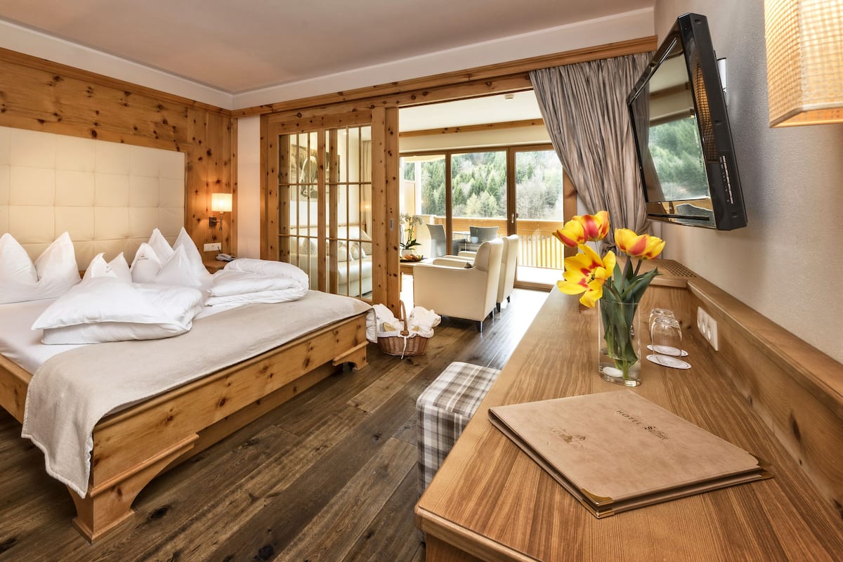 A wooden-floored bedroom with a large bed, a desk with a vase of flowers, and a view of the outdoors through large windows.