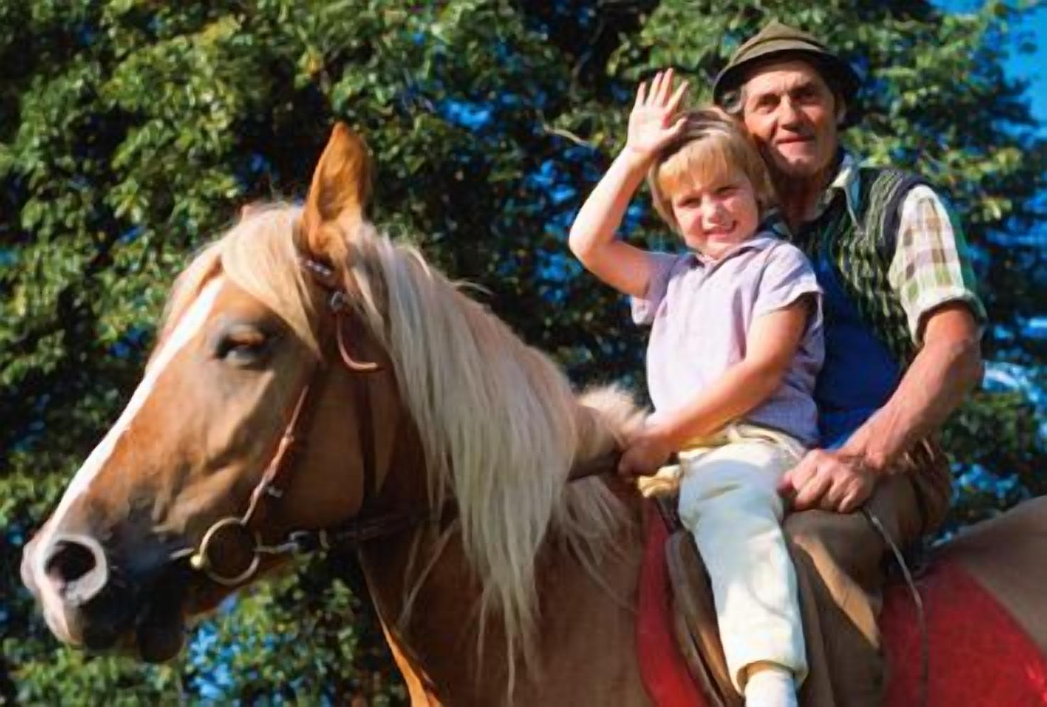 A person riding a horse with a child on the back, both wearing casual clothing.