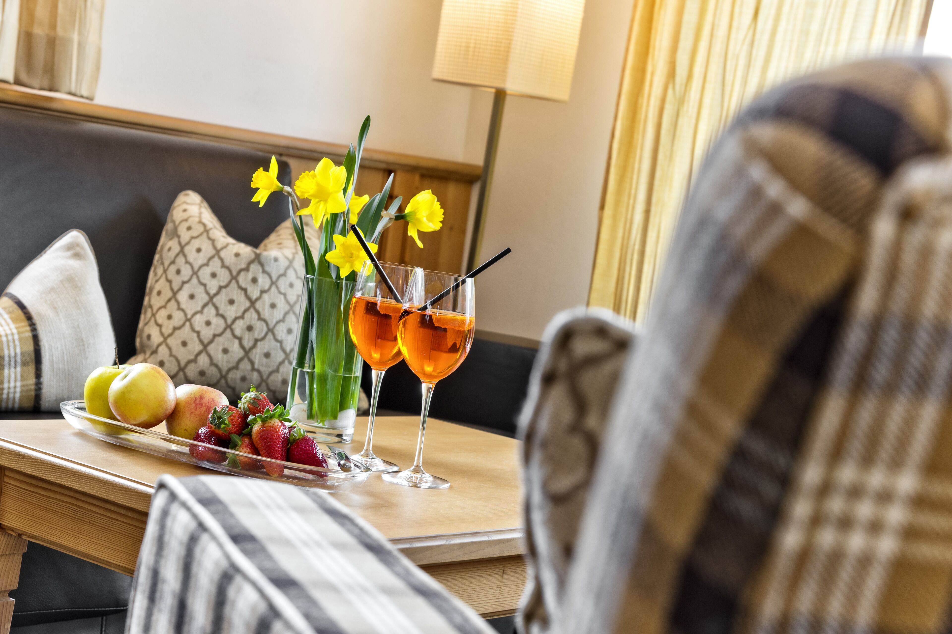 Une table à manger avec un plateau de fruits, deux verres de jus d’orange et un vase rempli de fleurs jaunes.
