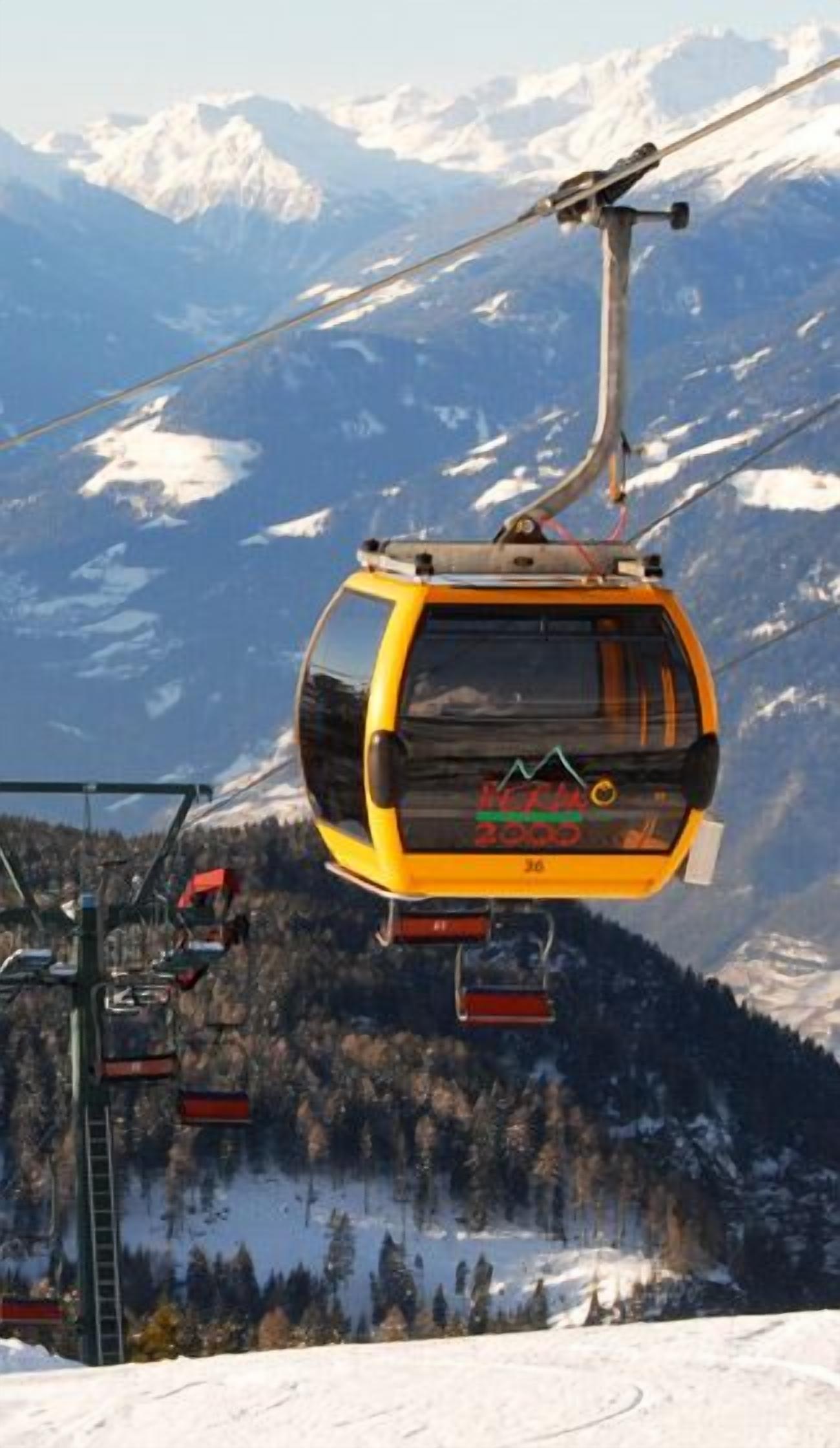 A yellow cable car with "MICHAEL 2000" on the front, suspended by cables against a backdrop of snow-covered mountains and trees.