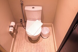 Un baño pequeño con un inodoro, un rollo de papel higiénico, un cabezal de ducha y un cesto de basura.