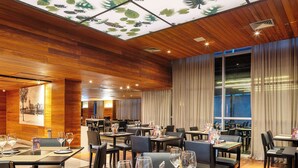 A restaurant with wooden panel ceilings, tables set with wine glasses and menus, and a view of the city through large windows. - Hotel Park Tower Savassi (Belo Horizonte)