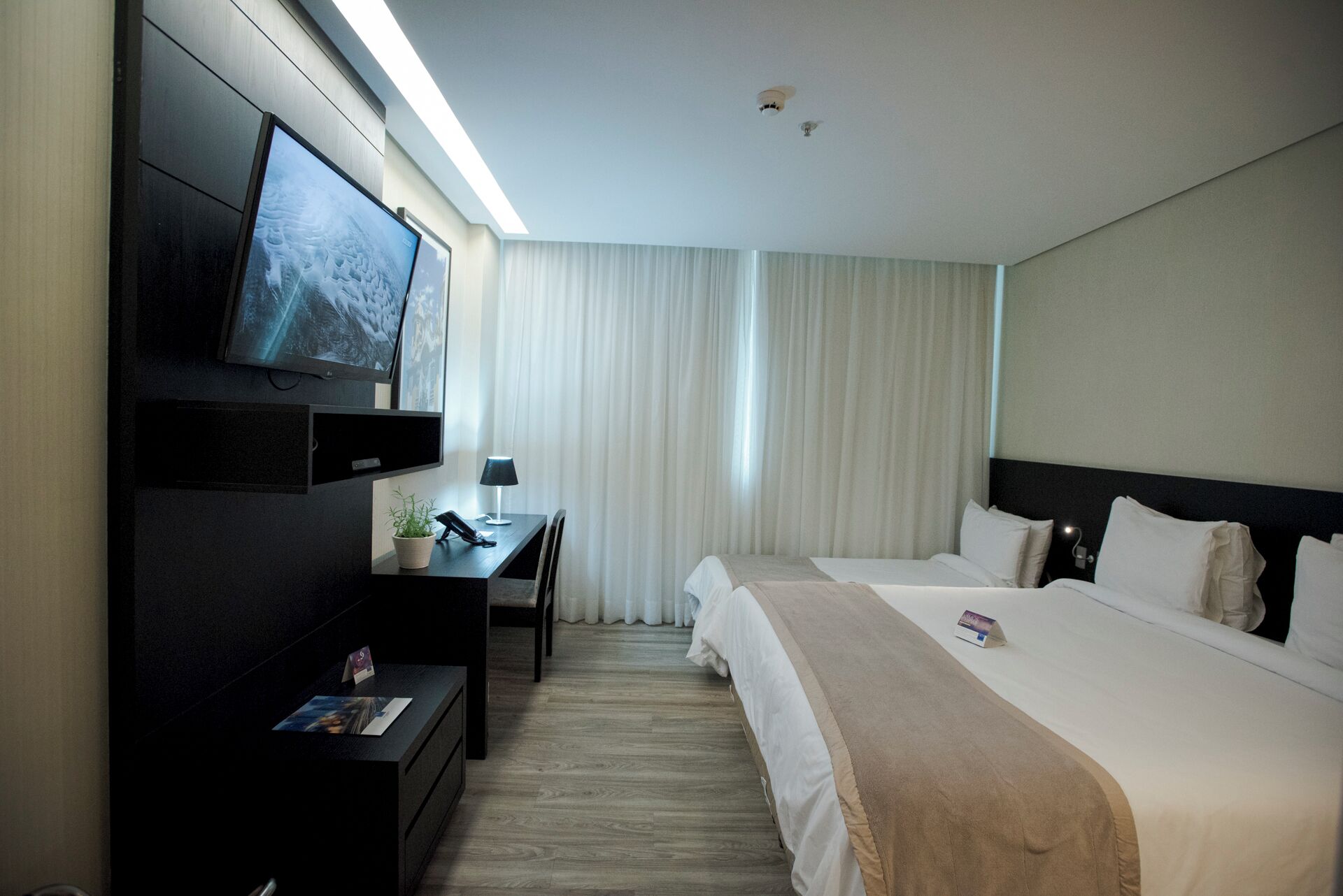 A hotel room with a bed, a desk with a lamp, a TV mounted on the wall, and a window with curtains.