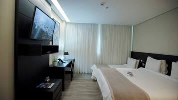 A hotel room with a bed, a desk with a lamp, a TV mounted on the wall, and a window with curtains.
