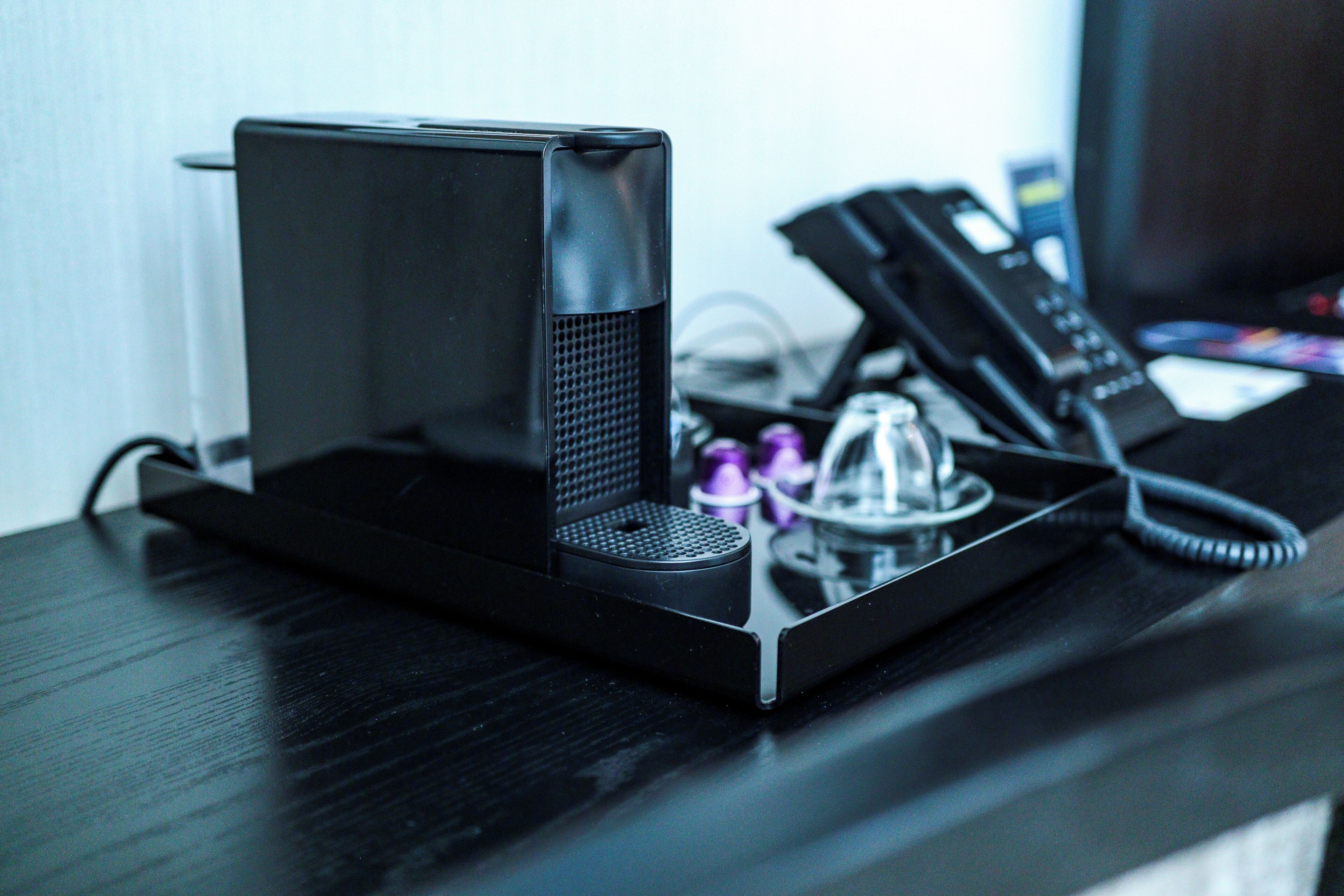 A black portable speaker on a desk with a telephone and a glass object.