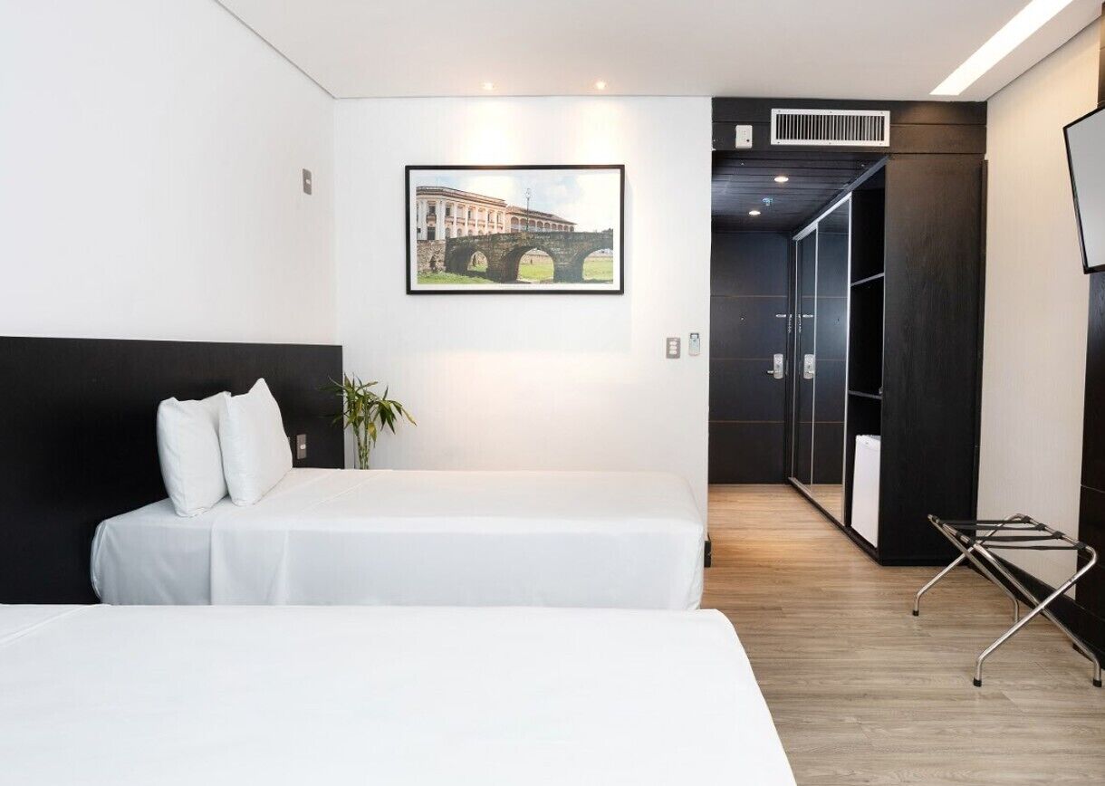 A modern hotel room with a large bed, a black headboard, a framed picture of a bridge, and a wooden floor.